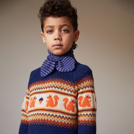 A boy in a striped blue neck tie wears an indigo Oeuf Kid's Fairisle Sweater with squirrels on it in front of a neutral background. 