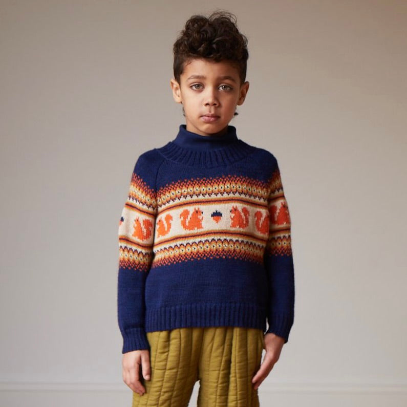 A boy wears an indigo Oeuf Kid's Fairisle Sweater with squirrels on it in front of a neutral background.