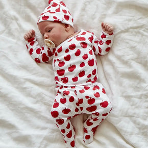 Take Me Home Outfit - baby in a white kimono style top, footie pants, and hat with red apple pattern on a neutral background