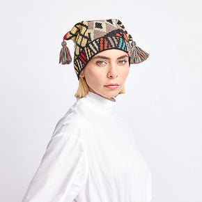 Kuna Watanabe Hat - model wearing yellow, black, red, and blue patterned hat with pom pom detail on a neutral background