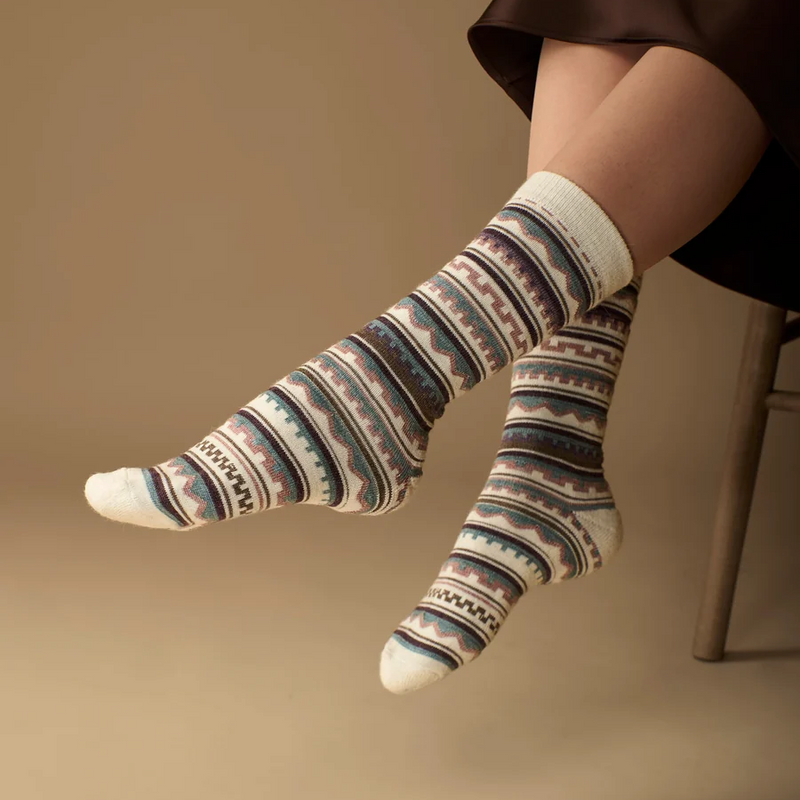 A person seated on a chair wearing patterned crew socks with horizontal bands in cream, blue, purple, and pink, against a warm beige studio background.