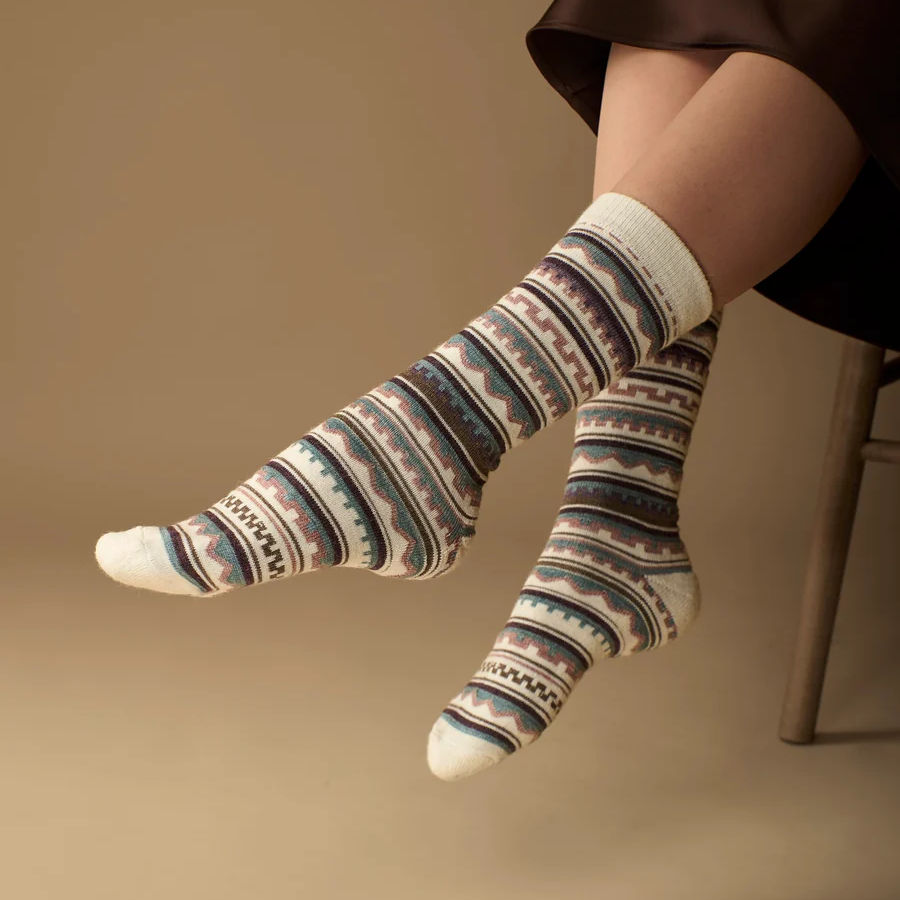 A person seated on a chair wearing patterned crew socks with horizontal bands in cream, blue, purple, and pink, against a warm beige studio background.
