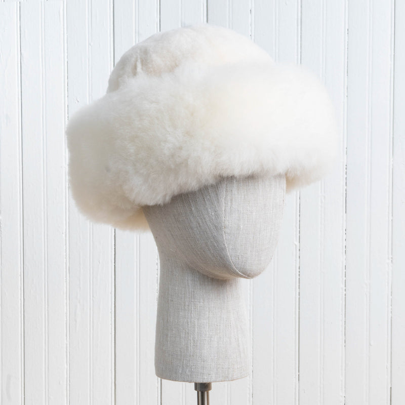 Premium Baby Alpaca Fur Hat. A natural white Premium Baby Alpaca Fur Hat on a mannequin head against a white paneled background.