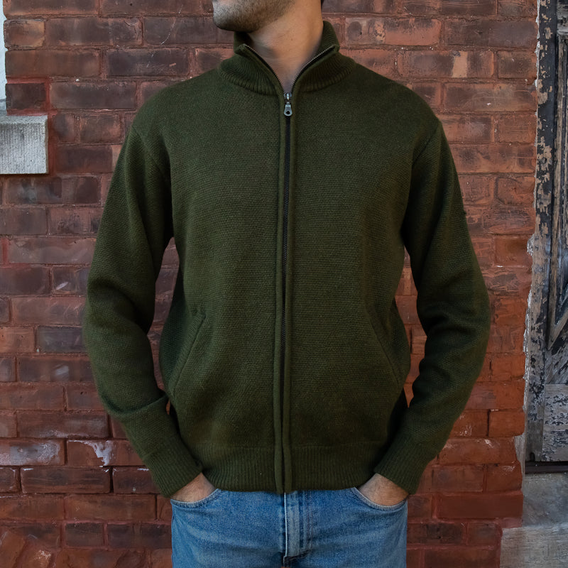 Men's Heavyweight Full-Zip 100% Baby Alpaca Wool Jacket. A man in an olive green knitted jacket stands in front of a brick wall. 