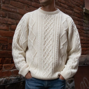 Alpaca Aran Cableknit Sweater. Man wearing a cream aran knit sweater against a brick wall. 