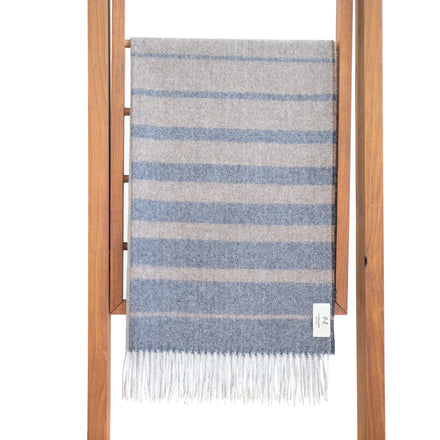 A striped baby alpaca shawl in gray and blue, displayed on a wooden hanger with fringe visible at the bottom.