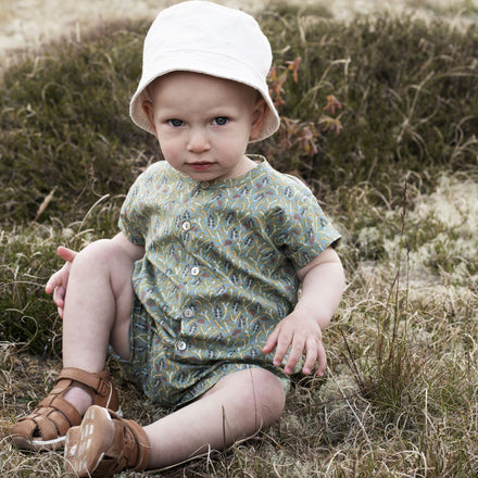 Serendipity Baby Button Suit - 100% GOTS Cotton, a  baby in a white hat wearing a green patterned button onesie agains, sitting in a field