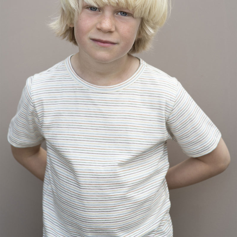 Serendipity Kid's Jersey Tee - 100% Organic Cotton, a blond child wearing a striped rainbow jersey tee against a grey background. 