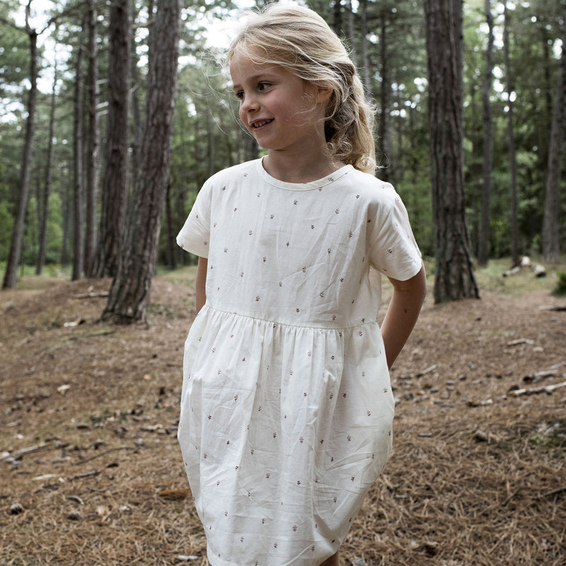 Serendipity Kid's Loose Dress - 100% Organic Cotton, front view of a girl wearing a White dress with reddish-brownish flowers in a forest