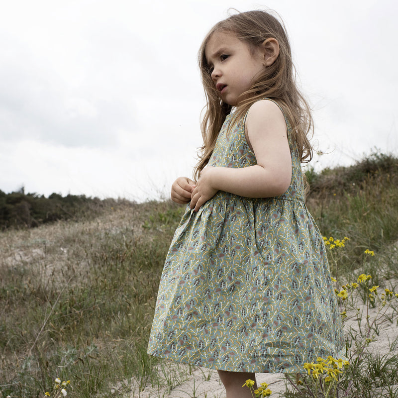 Serendipity Kid's Peasant Dress - 100% Organic Cotton | Fluff Alpaca, a child wearing a green peasant dress in field with yellow flowers