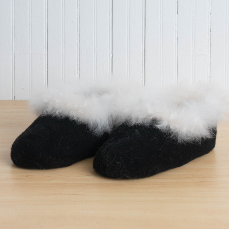 Black slippers with white fur trim on a wooden floor against a white paneled wall.