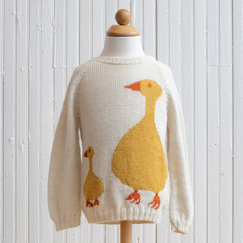 A beige baby alpaca sweater with a duck design on it, displayed on a wooden mannequin against a white wooden background.