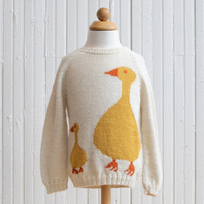 A beige baby alpaca sweater with a duck design on it, displayed on a wooden mannequin against a white wooden background.