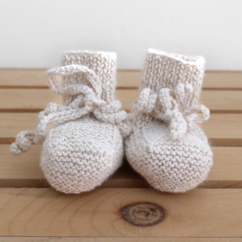 A pair of hand-knit alpaca baby booties in beige, with adjustable ties, displayed on a wooden surface.