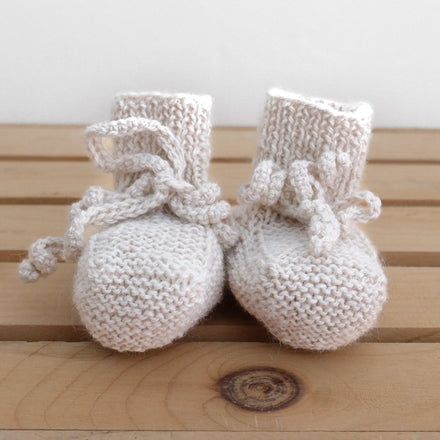 A pair of hand-knit alpaca baby booties in beige, with adjustable ties, displayed on a wooden surface.