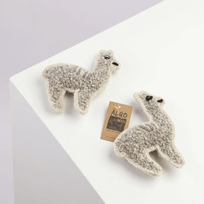 Two alpaca-shaped dog toys made of sheep's wool, displayed on a white surface with a product tag in the background.