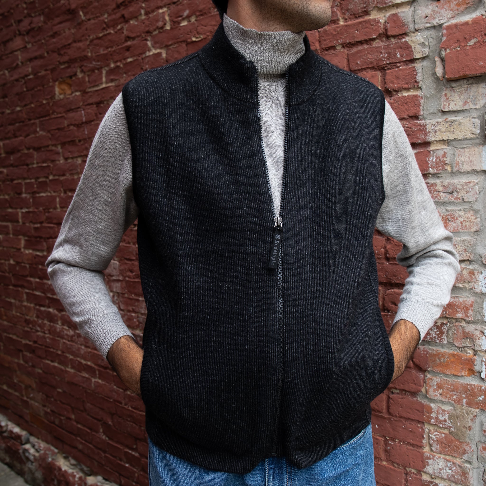 A man wearing a black Bucksport Zip Alpaca Vest over a grey sweater, with blue jeans and standing against a brick wall.
