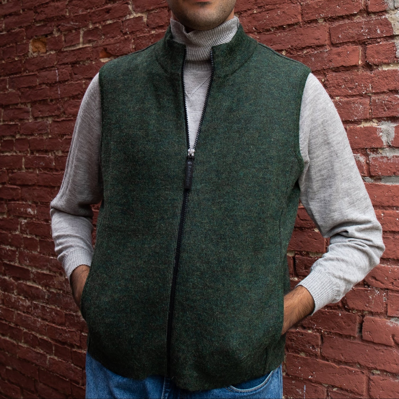 Person wearing a green vest over a gray sweater against a brick wall.