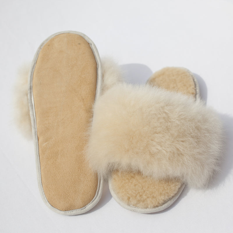 A pair of beige alpaca fur slides with a plush upper sole and cotton twill outer edge, displayed on a white background.