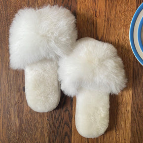 White fluffy slippers on a wooden surface with a plate of cake and a cup in the background.