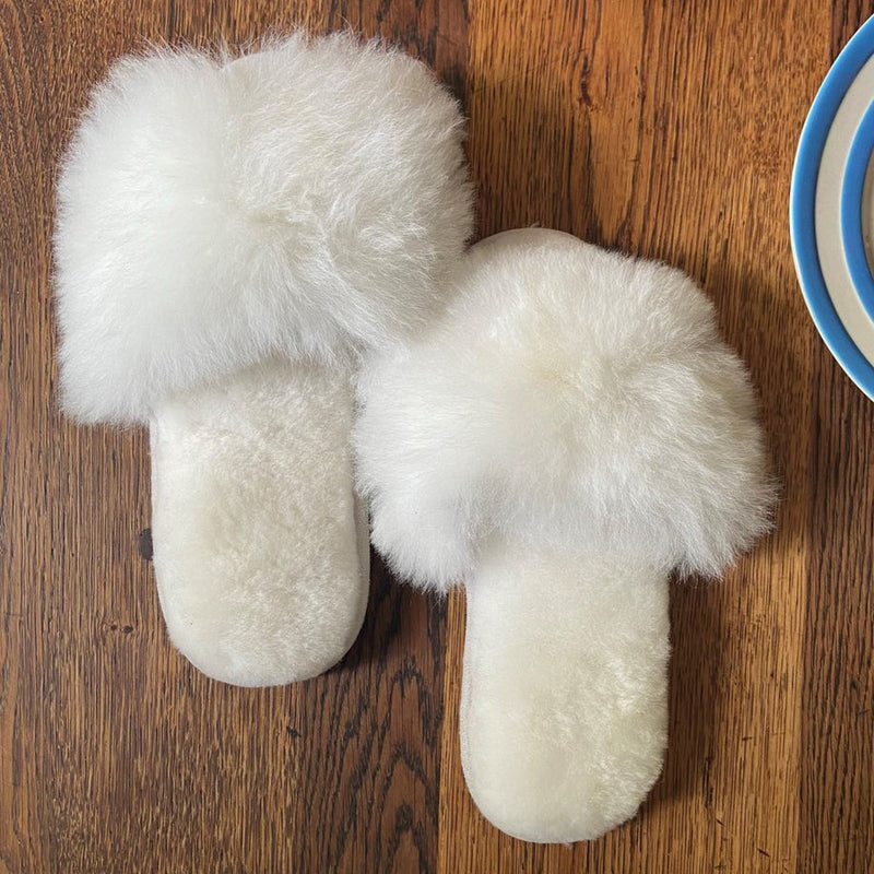 White fluffy slippers on a wooden surface with a plate of cake and a cup in the background.